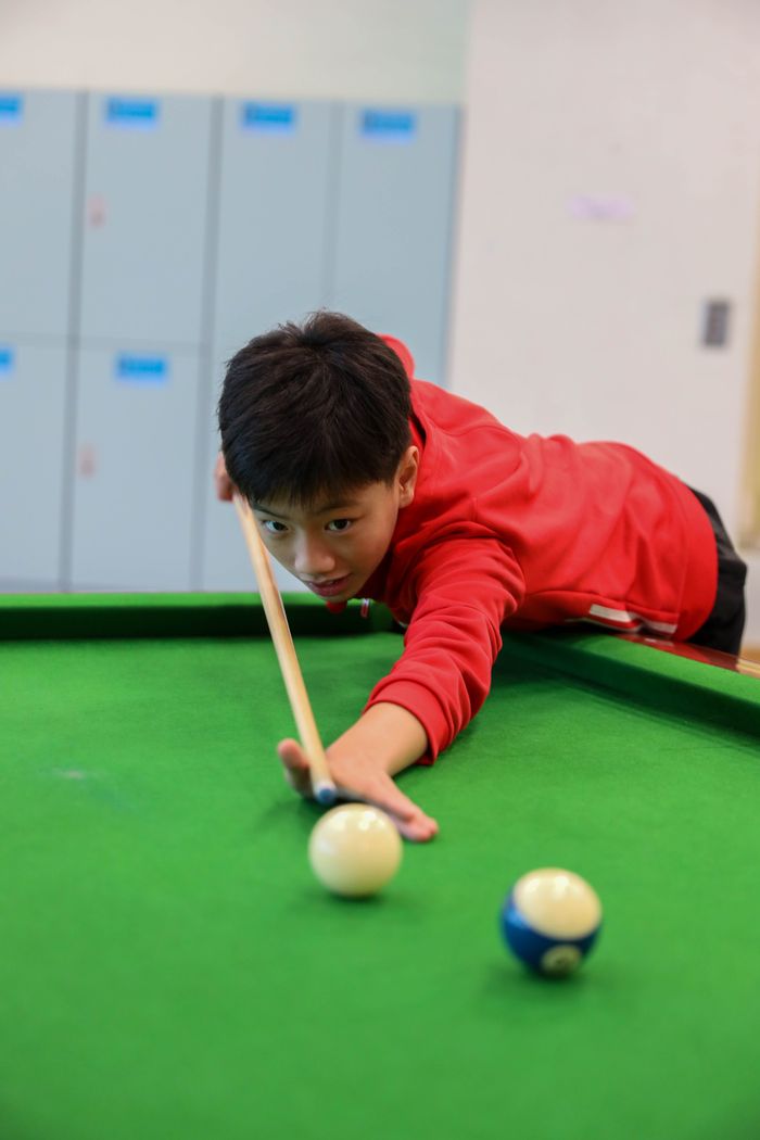 A young boy in red shirt aims carefully at a snooker table, concentrating intensely on his shot.
