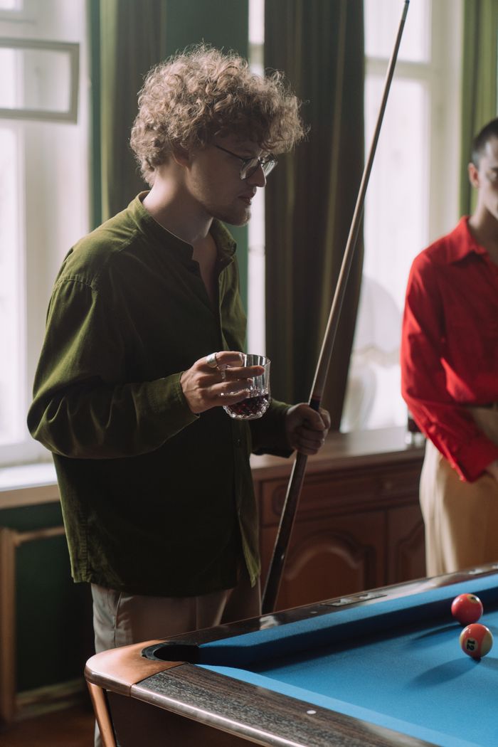 A young man with curly hair holds a cue stick and whiskey while playing billiards indoors.